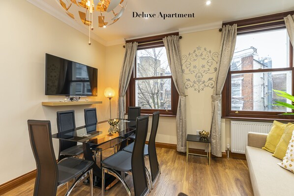 A modern living room with a dining area, a flat-screen TV mounted on the wall, and a sofa with cushions. - Kensington Olympia (London)