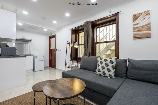 A modern living room with a grey sofa, two round wooden tables, and a kitchen area with a refrigerator and microwave.