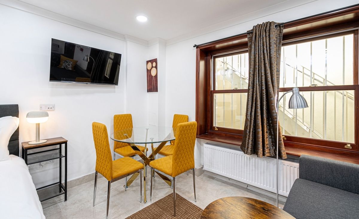 A hotel room with a bed, a TV mounted on the wall, a dining table with chairs, a window with curtains, and a radiator.