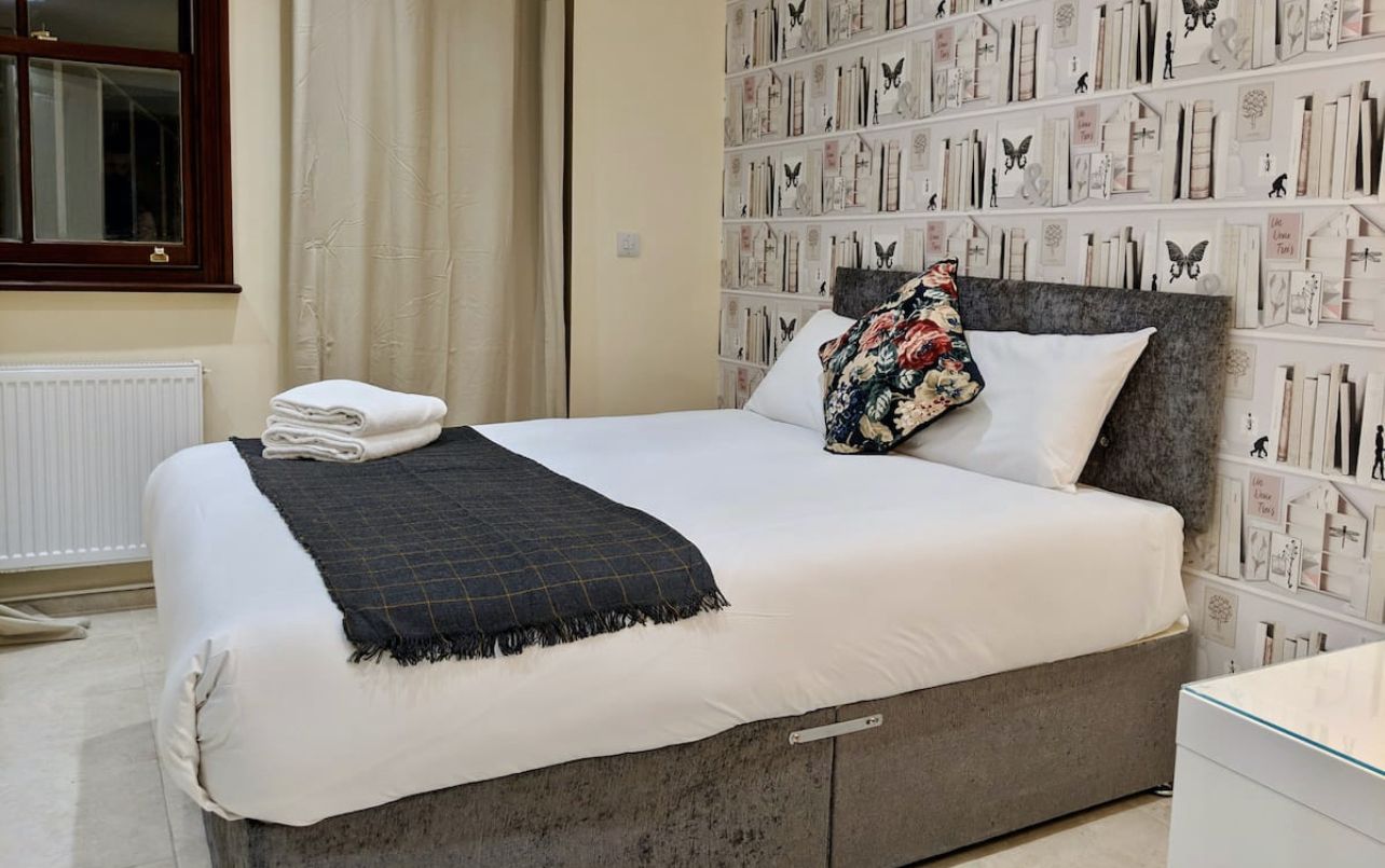 Una cama bien hecha con un edredón blanco y una manta de cuadros oscuros, un cojín floral y una ventana con cortinas.