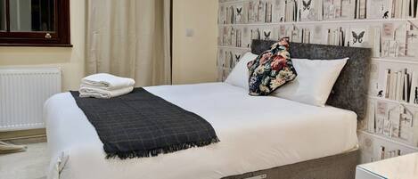 Una cama bien hecha con un edredón blanco y una manta de cuadros oscuros, un cojín floral y una ventana con cortinas.