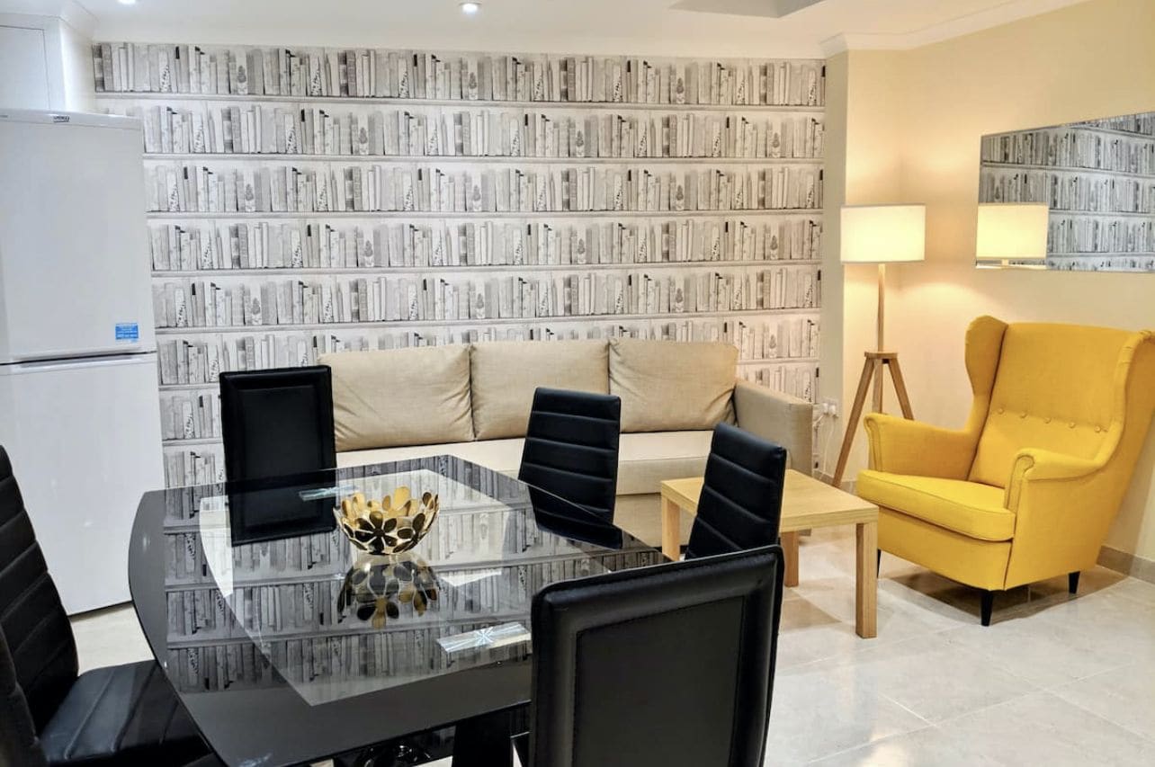 A modern living room with a glass-top dining table, black chairs, a yellow armchair, a sofa, and a bookshelf in the background.