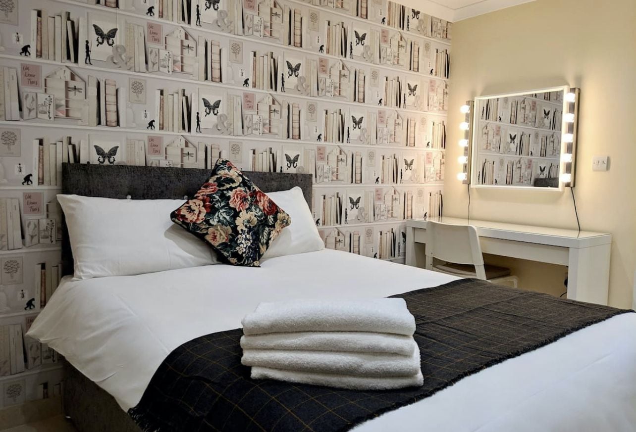 A neatly made bed with a floral pillow and a stack of towels, a bedside table, and a wall with a mirror and lights.