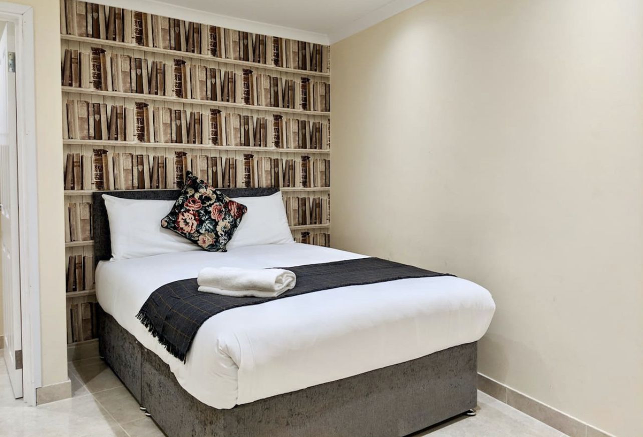 A neatly made bed with a white comforter and a dark blanket, a pillow with a floral pattern, and a folded towel.