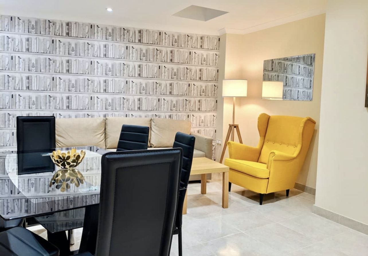 A modern living room with a glass coffee table, a sofa, a yellow armchair, and a bookshelf against the wall.