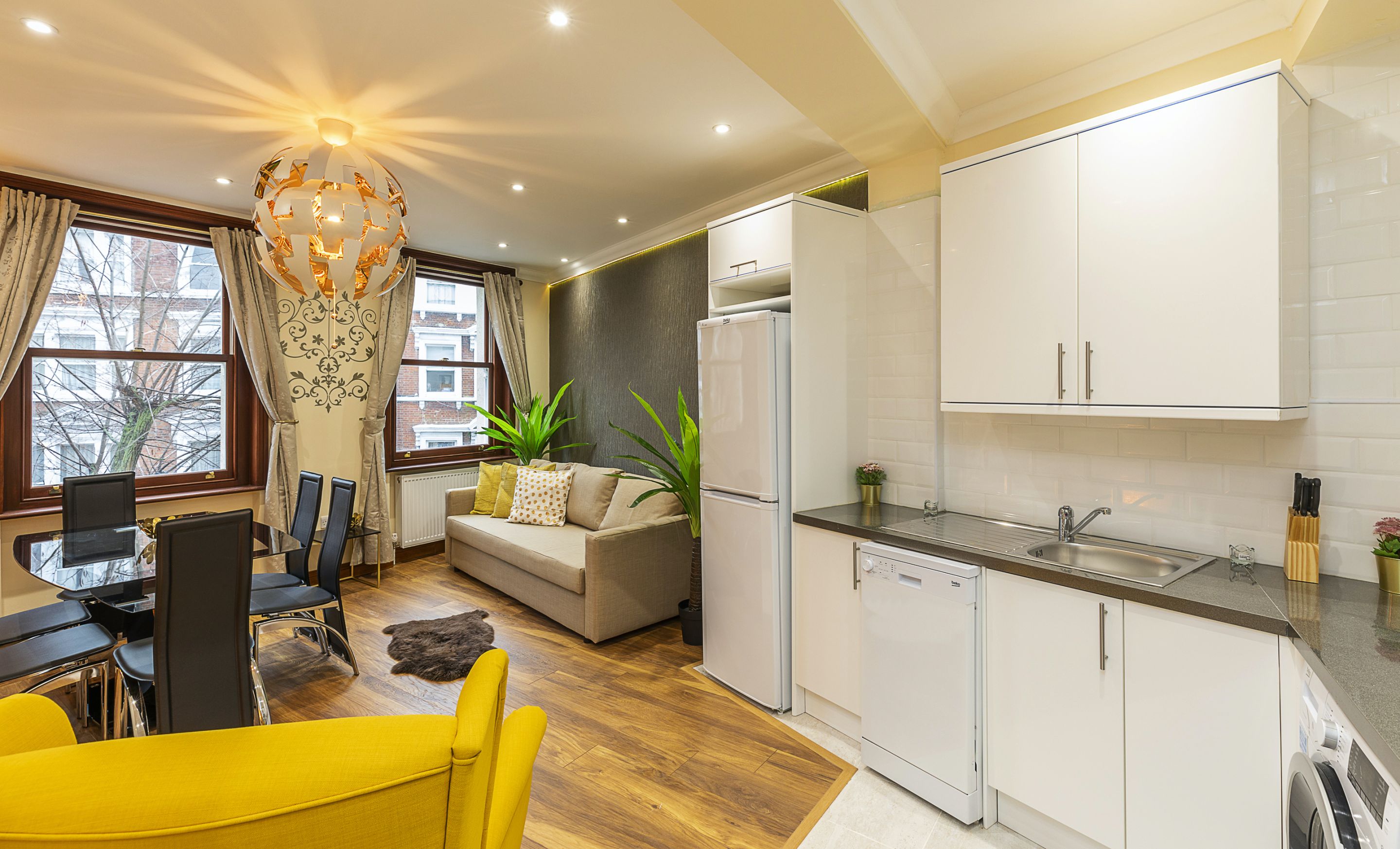 A modern kitchen with white cabinets, a built-in oven, and a sink. There is a dining area with a yellow table and chairs, and a sofa.