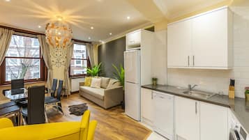 A modern kitchen with white cabinets, a built-in oven, and a sink. There is a dining area with a yellow table and chairs, and a sofa.