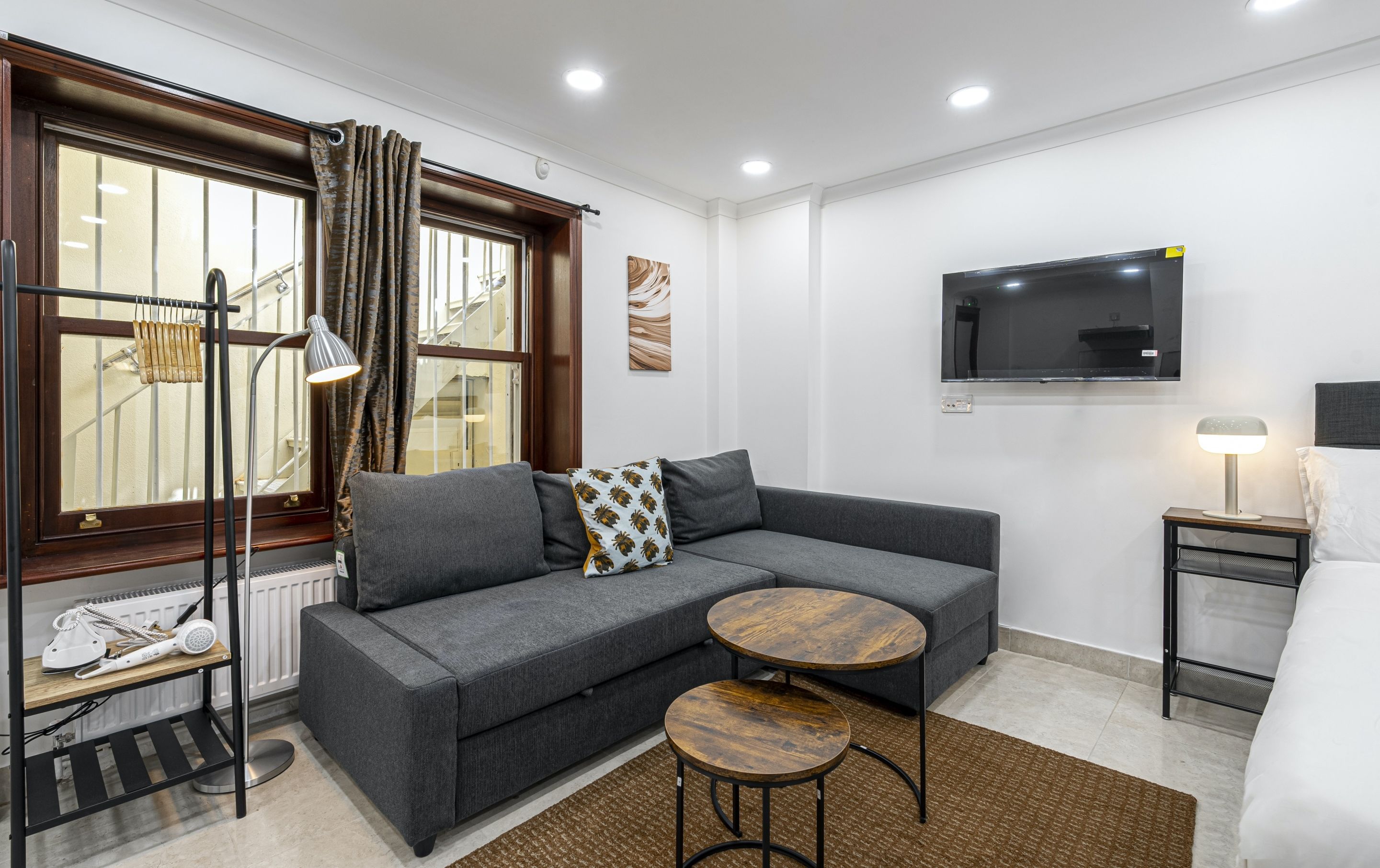 A modern hotel room with a grey sofa, a small wooden table, a TV mounted on the wall, and a bedside table with a lamp.