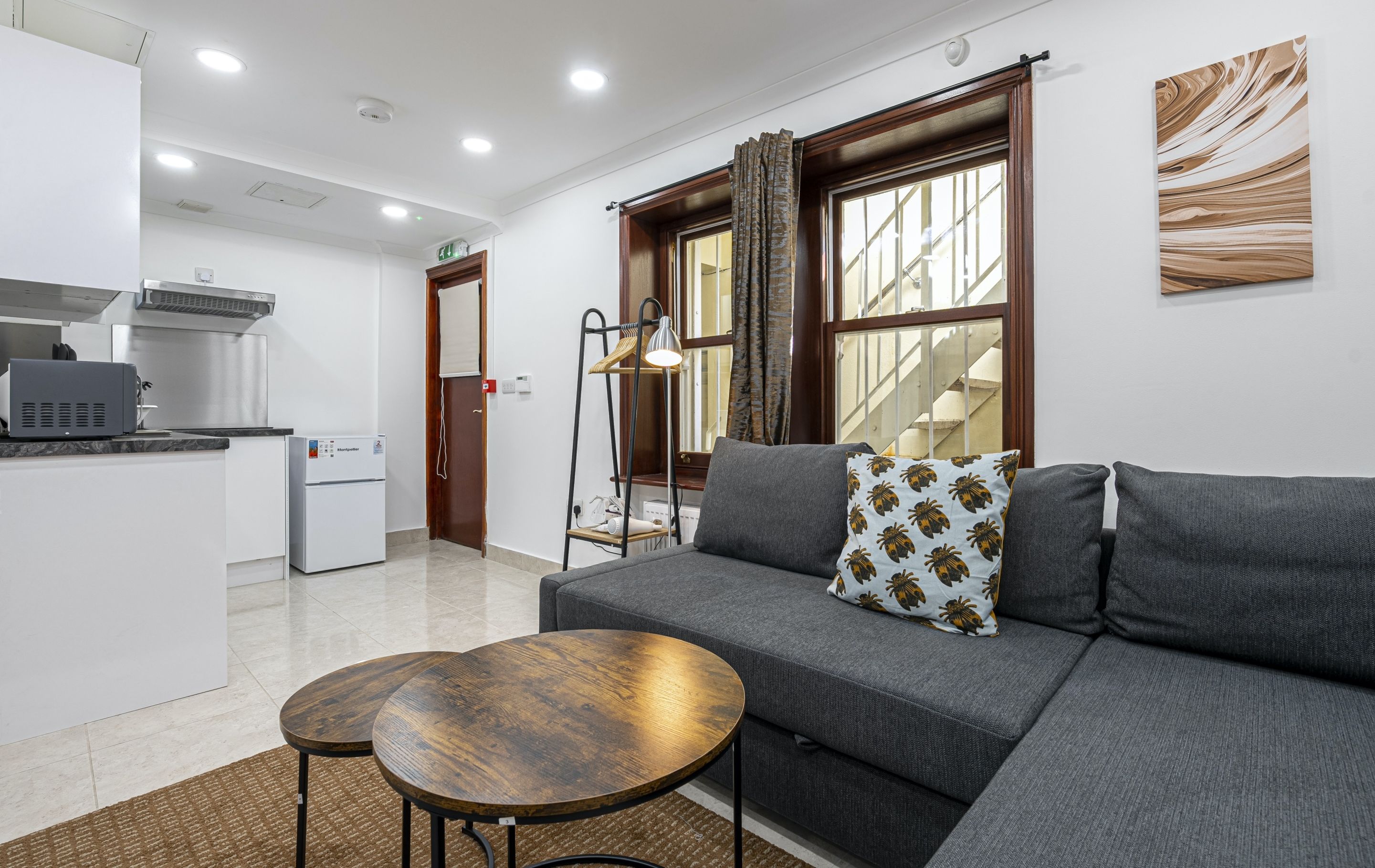 A modern living room with a grey sofa, two round wooden tables, and a kitchen area with a refrigerator and microwave.