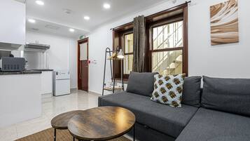 A modern living room with a grey sofa, two round wooden tables, and a kitchen area with a refrigerator and microwave.