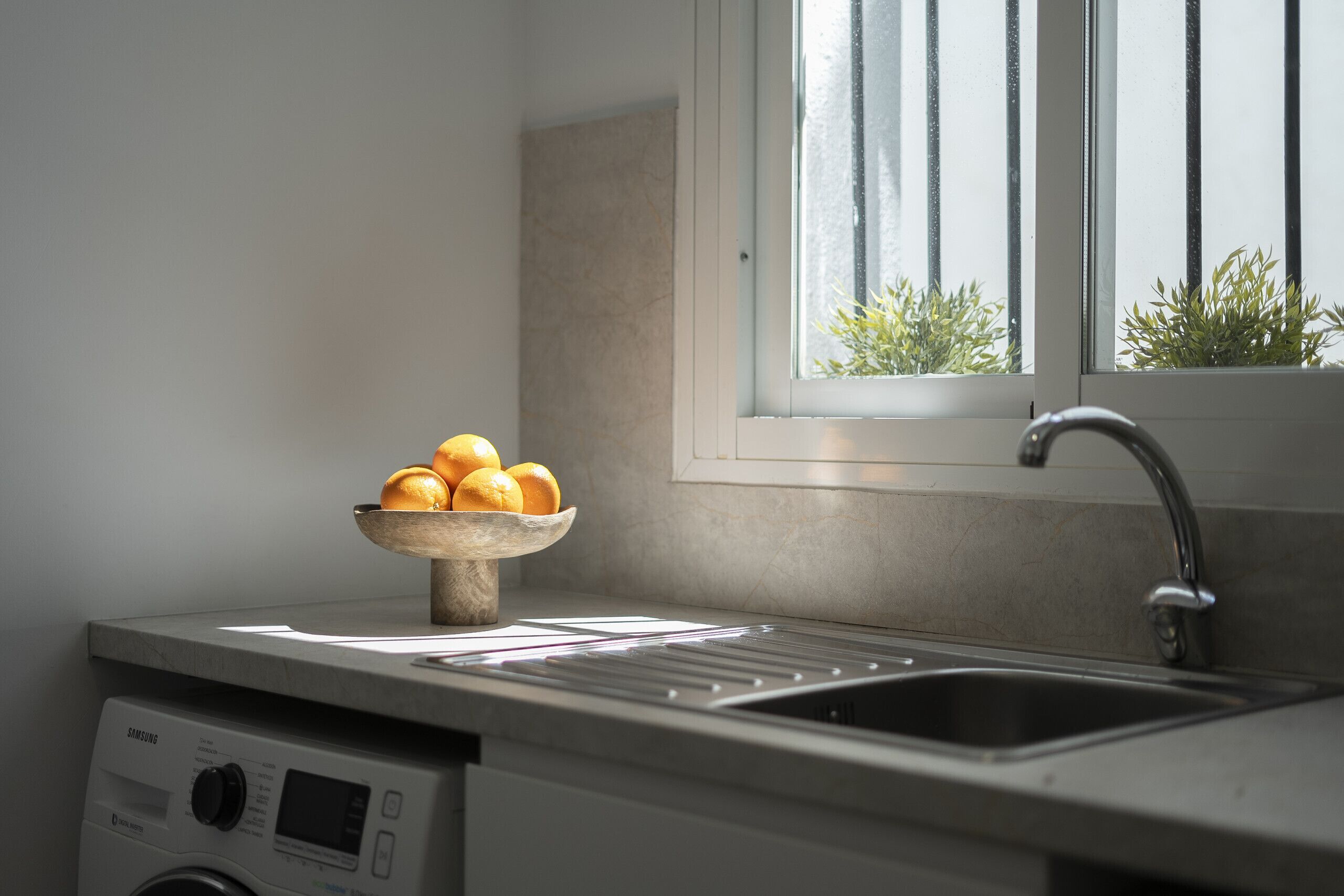 Een keuken met een spoelbak, een schaal oranje en een raam met planten.