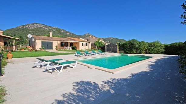 Un área de piscina con sillones de descanso, una casa con techo de tejas y montañas al fondo.