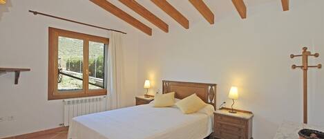 Un dormitorio con cama, lámparas de noche, ventana con vistas, radiador y techo de madera.