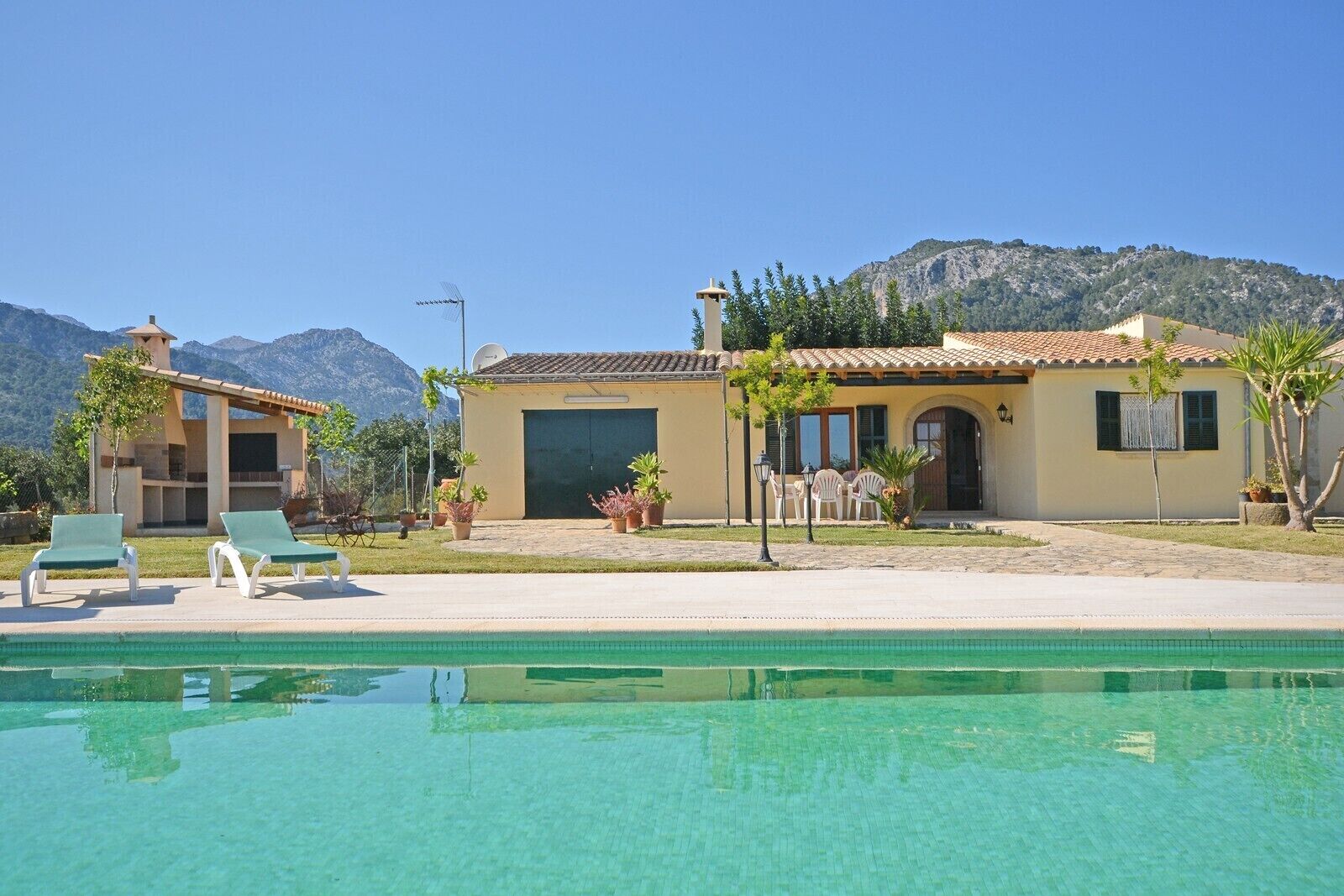 Un espace aménagé au bord de la piscine avec des chaises longues, un bâtiment doté d’un toit en tuiles et des montagnes en arrière-plan.
