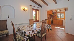 Un comedor con una mesa cubierta por un mantel con motivos de chocolate, sillas de madera y una cocina rústica al fondo.