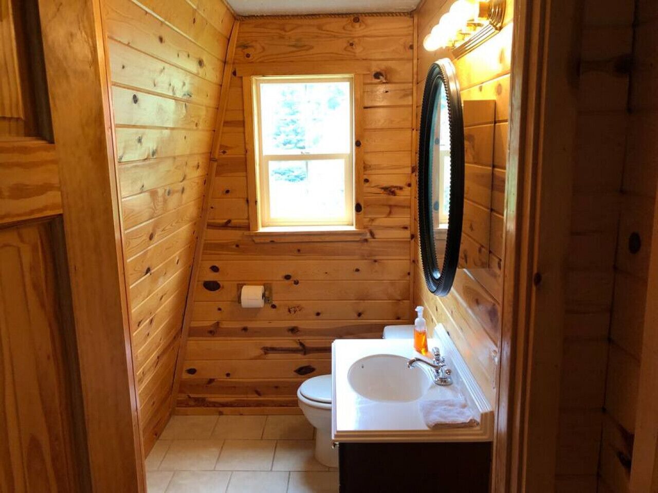 A small, wooden cabin bathroom with a window, toilet, and sink.