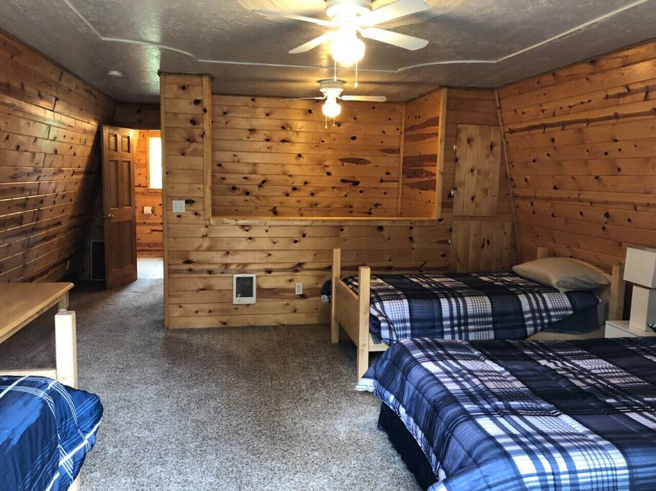 Une chambre de style cabane avec des murs en bois, deux lits avec une literie à motifs écossais, un ventilateur de plafond et une petite fenêtre.