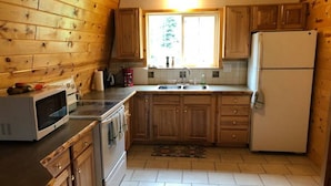 A compact kitchen with wooden cabinets, a microwave, a refrigerator, and a window providing natural light. - Balcony, 20 min to Yellowstone, Sled/ATV Spot (Island Park)
