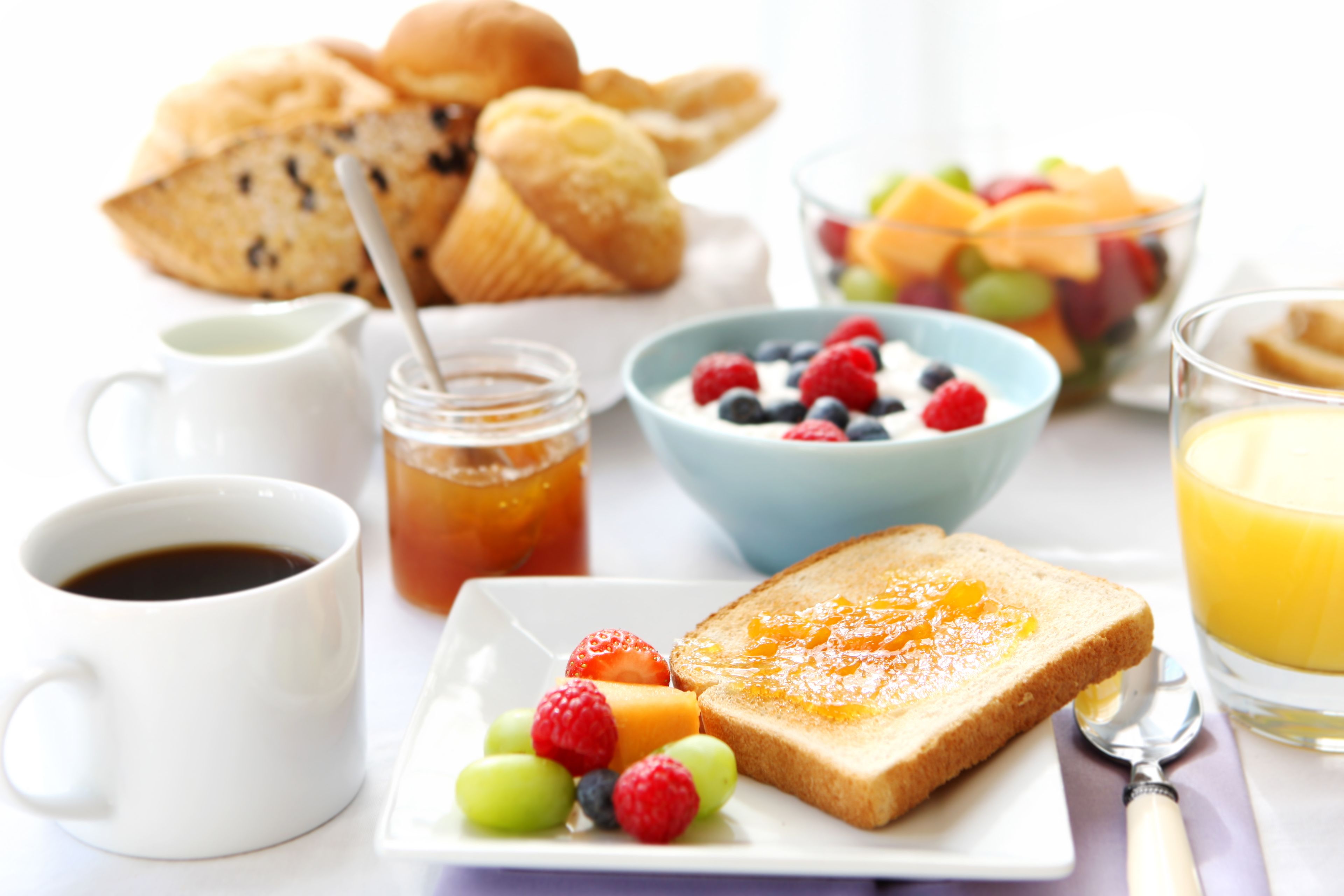 A breakfast table with toast, fruit, a bowl of cereal, a cup of coffee, a glass of orange juice, and a jar of jam.