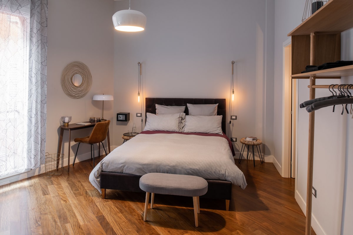 A bedroom with a bed, bedside tables, a desk, a chair, a hanging coat rack, and a window with curtains.
