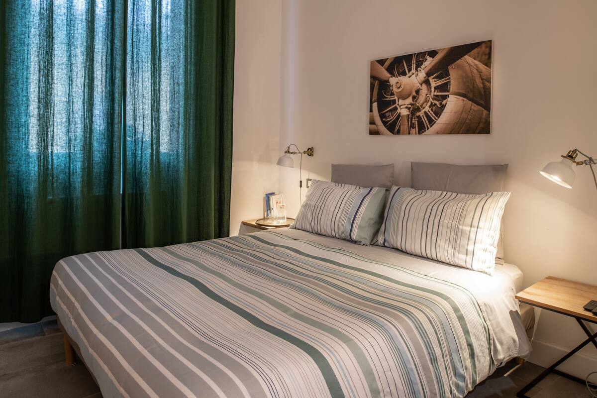 A neatly made bed with striped bedding, a wall-mounted clock, and a small bedside table with a lamp.