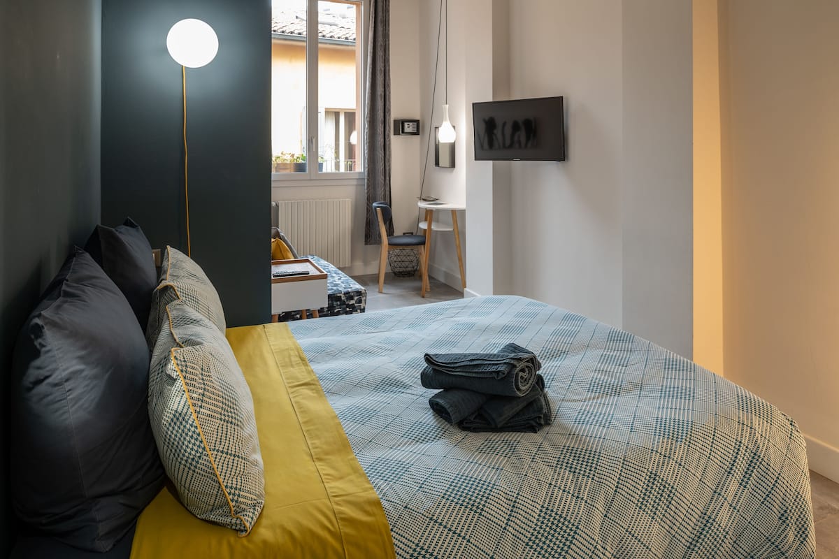A bedroom with a bed, pillows, a bedside table, a chair, and a view of the outside.