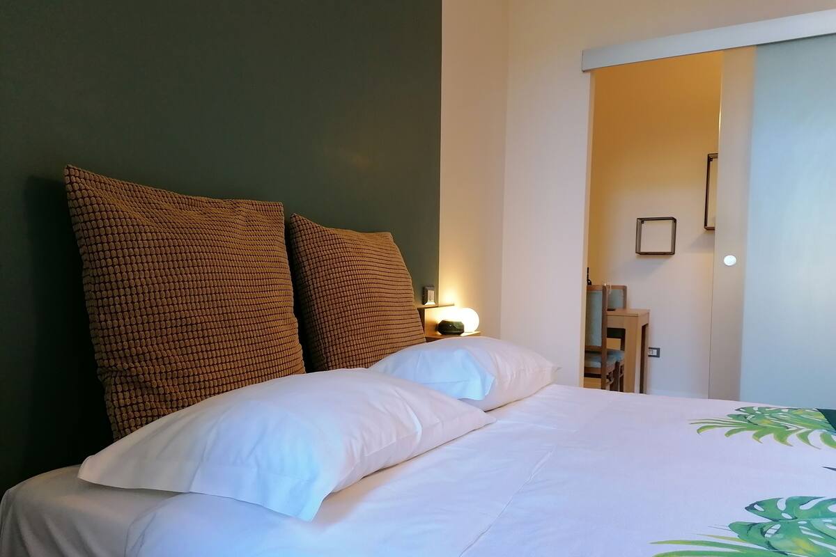 A neatly made bed with white bedding and two large brown pillows, a bedside table with a lamp, and a chair in the background.