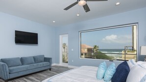 A bedroom with a large bed, a blue sofa, a TV mounted on the wall, and a large window with a view of the ocean. - Modern Beach House for 24! Private Pool, Ocean Views, & Directly Across from Beach! (South Padre Island)