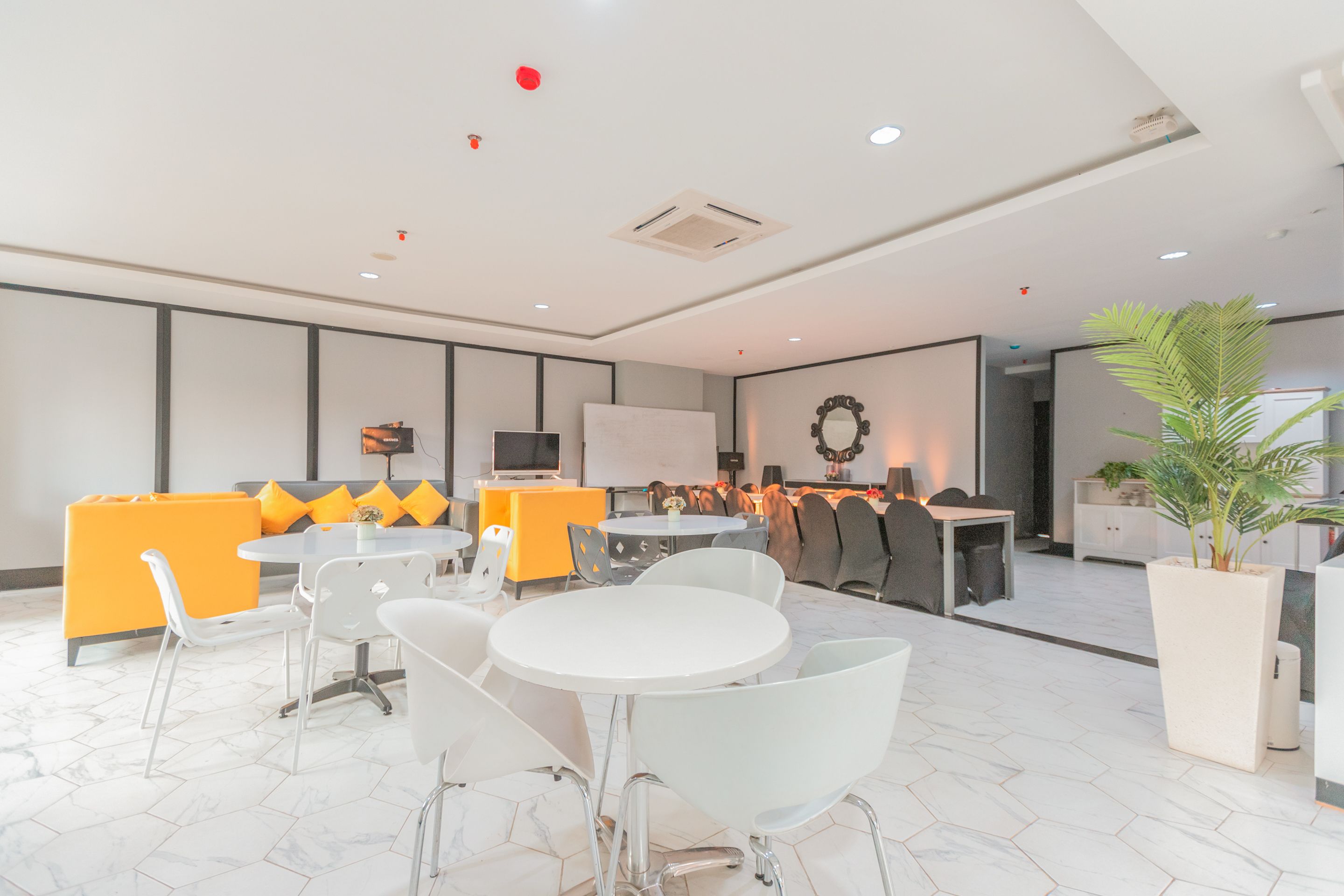 Un salón de conferencias moderno con mesas y sillas blancas, una pizarra grande y una zona de estar con cojines naranjas.
