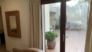 Una habitación con una cama de madera, un espejo y un ventanal corredera que da a un patio exterior con sillas y una planta en maceta.