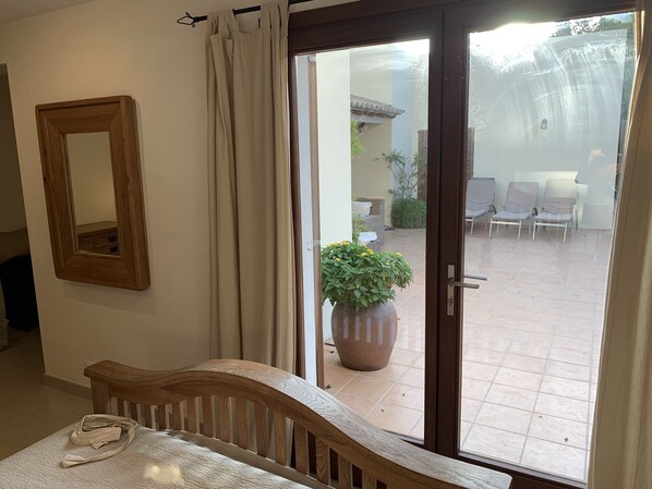 Una habitación con una cama de madera, un espejo y un ventanal corredera que da a un patio exterior con sillas y una planta en maceta.