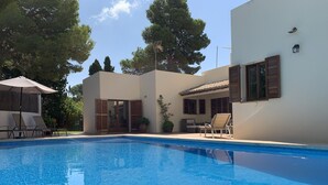 Piscina con sillones de descanso y zona cubierta, rodeada de árboles y un edificio con tejado de baldosas.