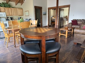 Un coin repas avec une table et des chaises en bois, un jeu d’échecs, et une cuisine en arrière-plan.