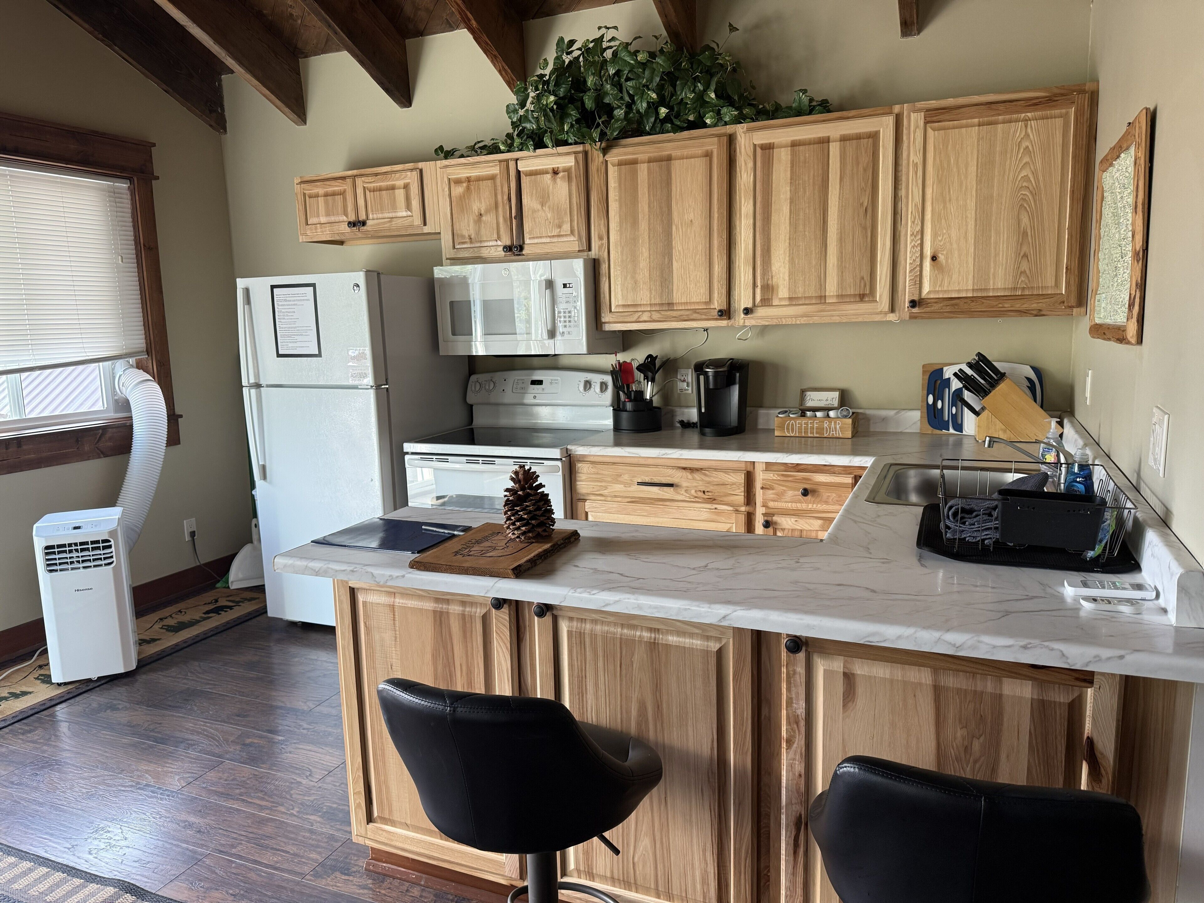 A kitchen with wooden cabinets, a white refrigerator, a microwave, a stove, and a coffee bar. There are two black bar stools and a potted plant on the counter.