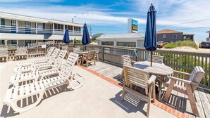 A poolside area with lounge chairs and umbrellas, a wooden deck, and a building in the background.