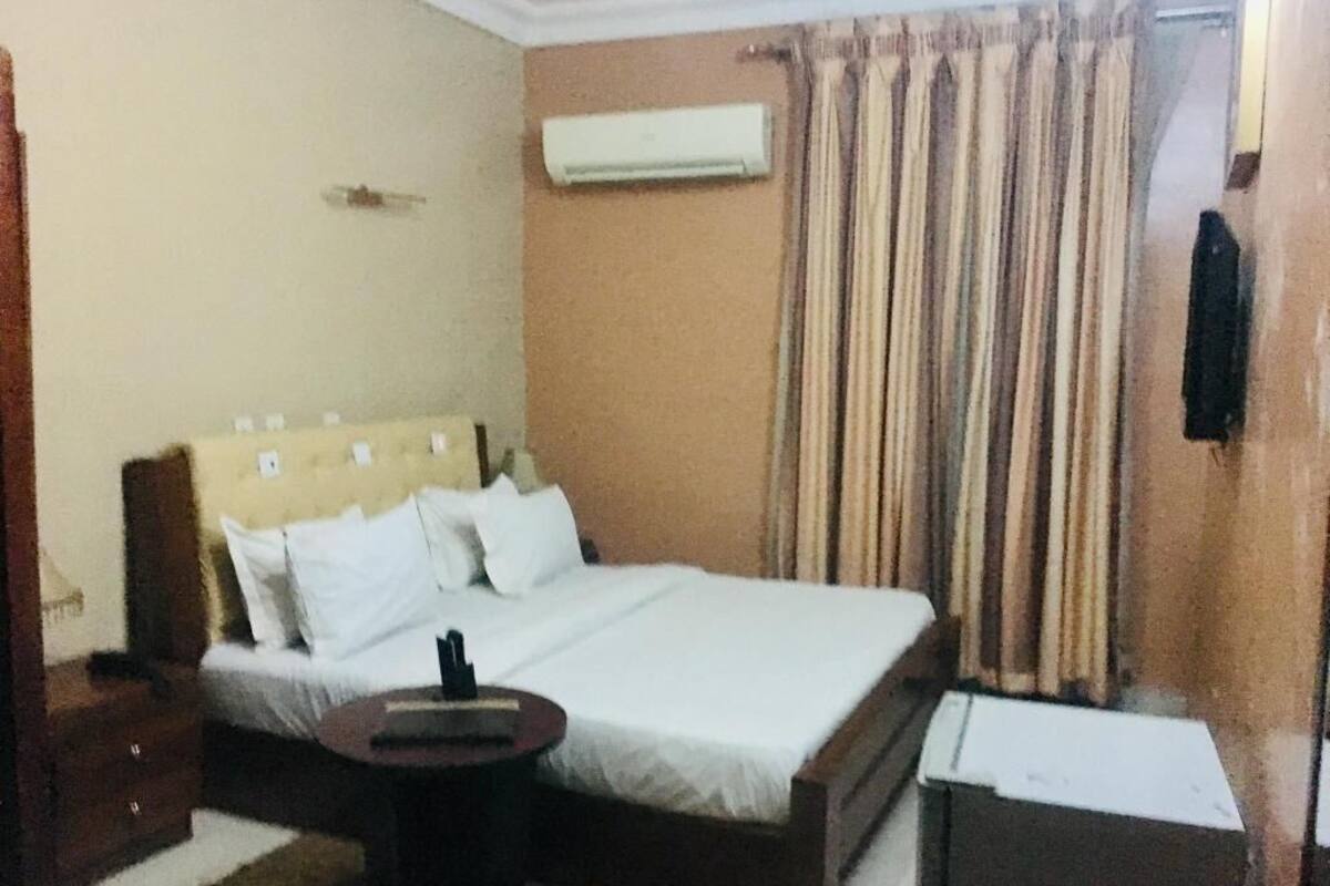 Habitación de hotel con cama, mesita de noche, aire acondicionado, cortinas y un pequeño escritorio.