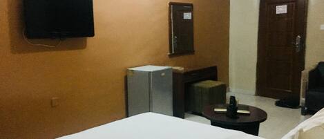 Habitación de hotel con cama, televisor montado en la pared, pequeño escritorio con silla, puerta y un mueble también sujeto a la pared.