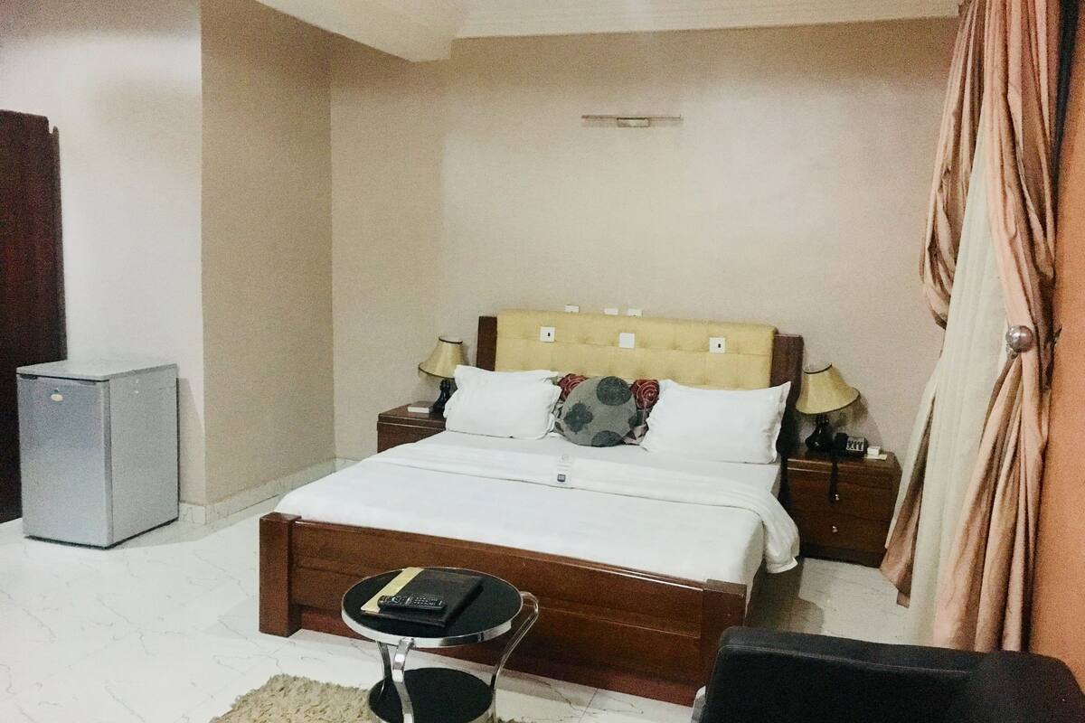 Un dormitorio con cama, mesitas de noche, una silla, una mesita pequeña y un refrigerador.