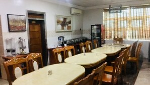 A dining area with round tables and wooden chairs, a kitchen area with appliances, and a window with blinds. - Euc Demodern Hotel & Suites (Lagos)