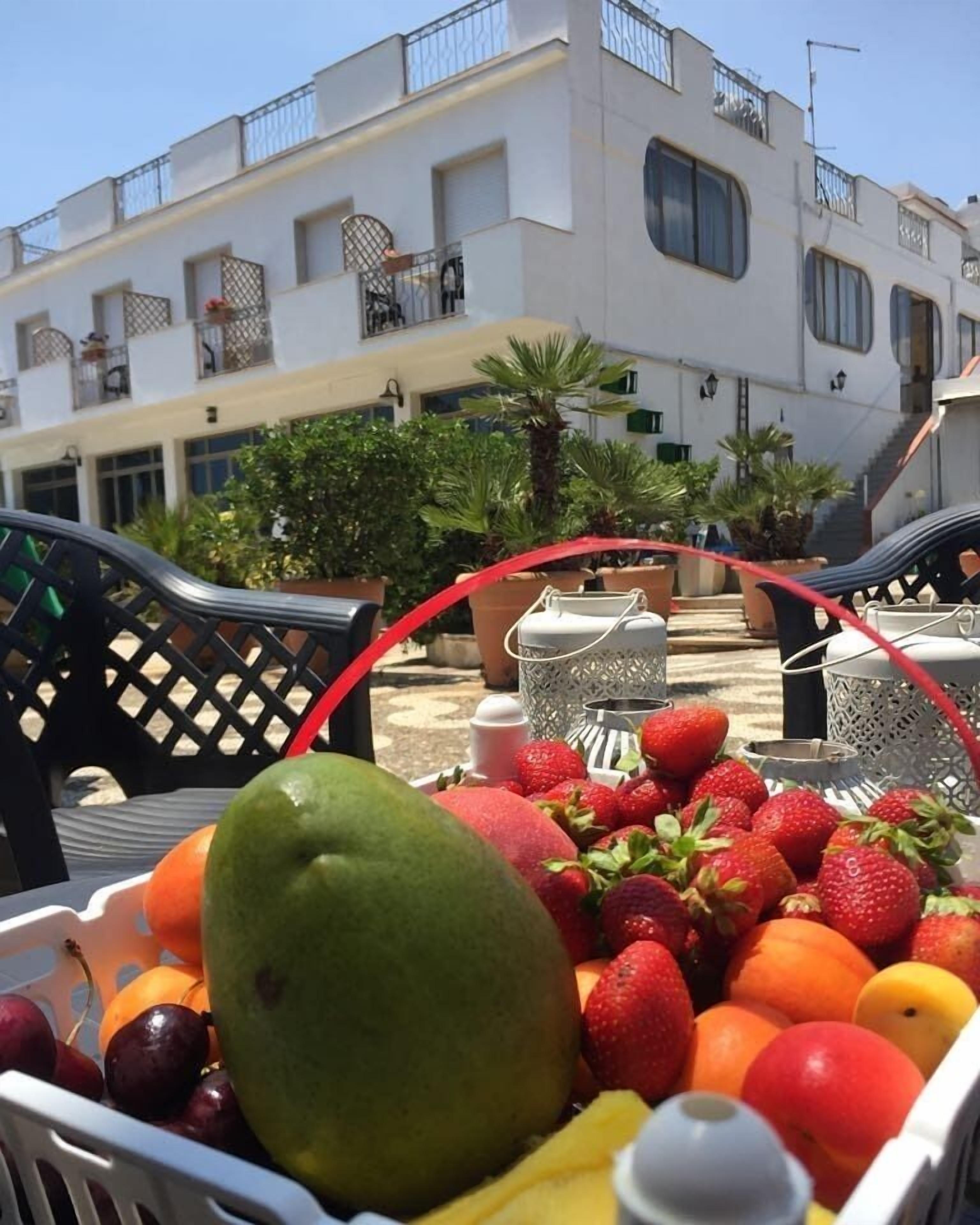 Een fruitmand met verschillende soorten fruit, zoals appels, sinaasappels en aardbeien, op een tafel voor een gebouw met balkons en bloempotten.