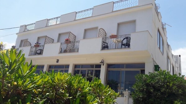 Un edificio blanco de varios pisos con balcones, ventanas amplias y algunas plantas en macetas.