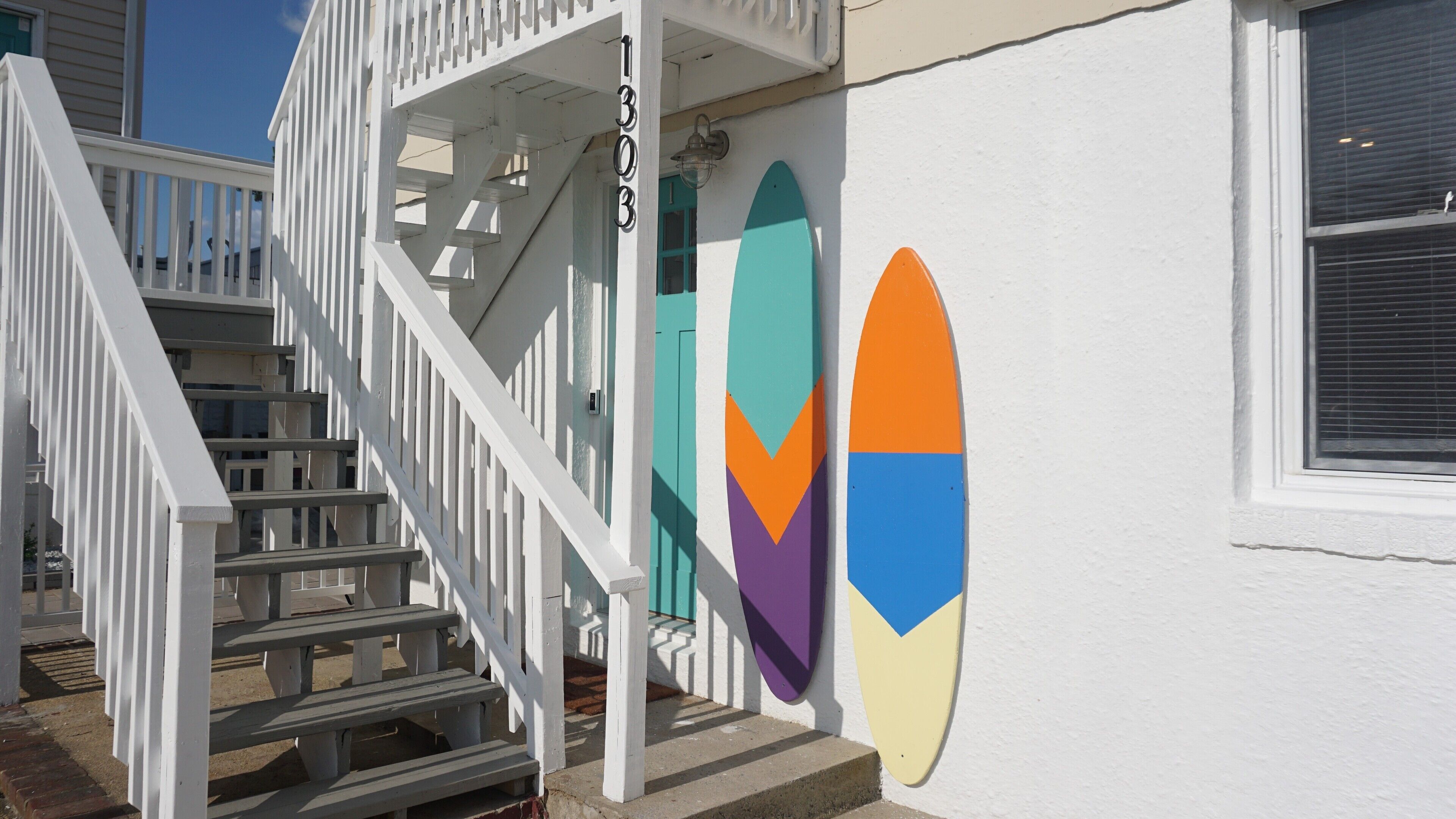 Un bâtiment blanc avec un escalier, une porte et trois planches de surf accrochées au mur.