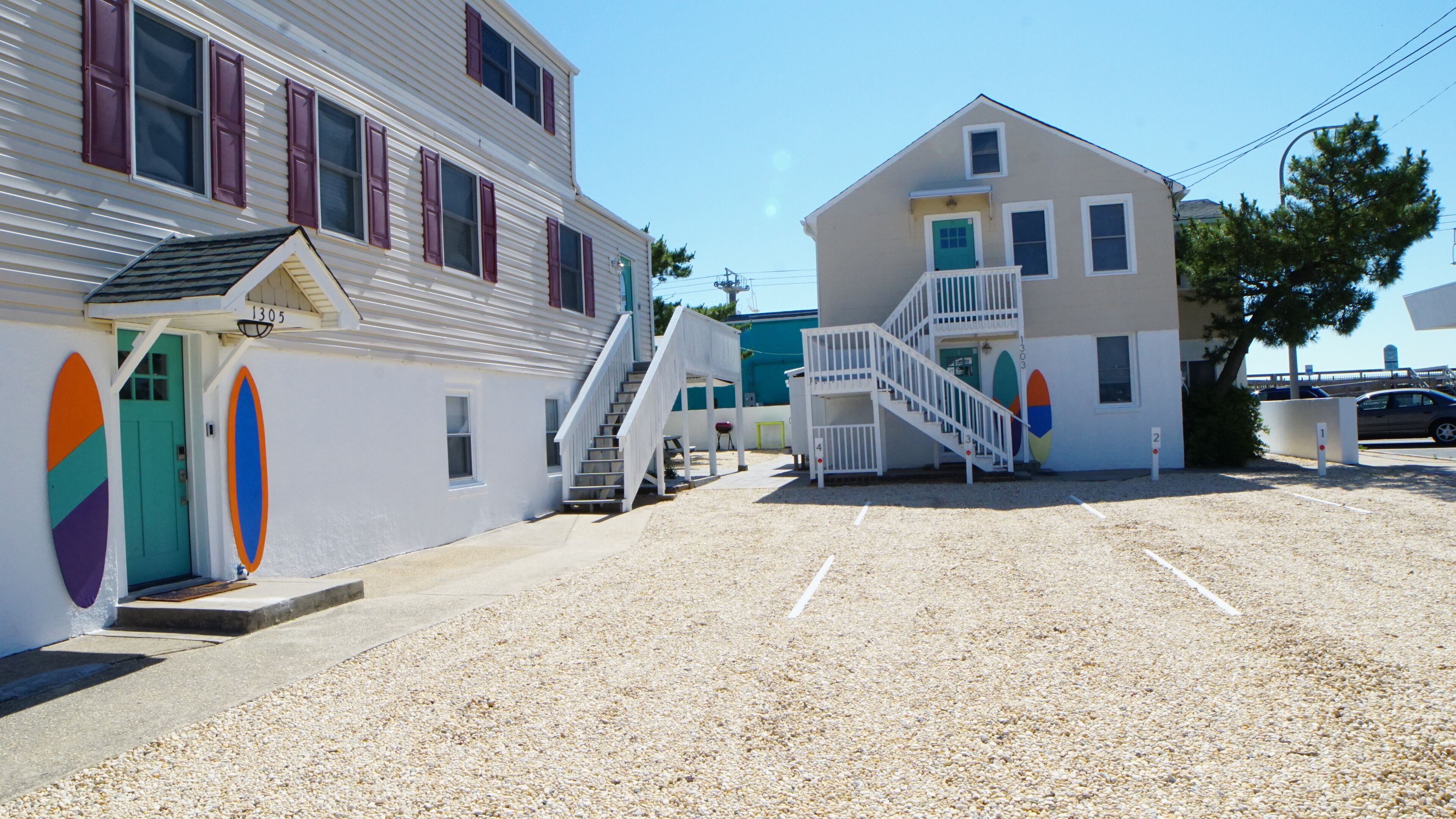 A building with a staircase leading to a second floor, a parking area with gravel, and colorful surfboard decorations on the walls.