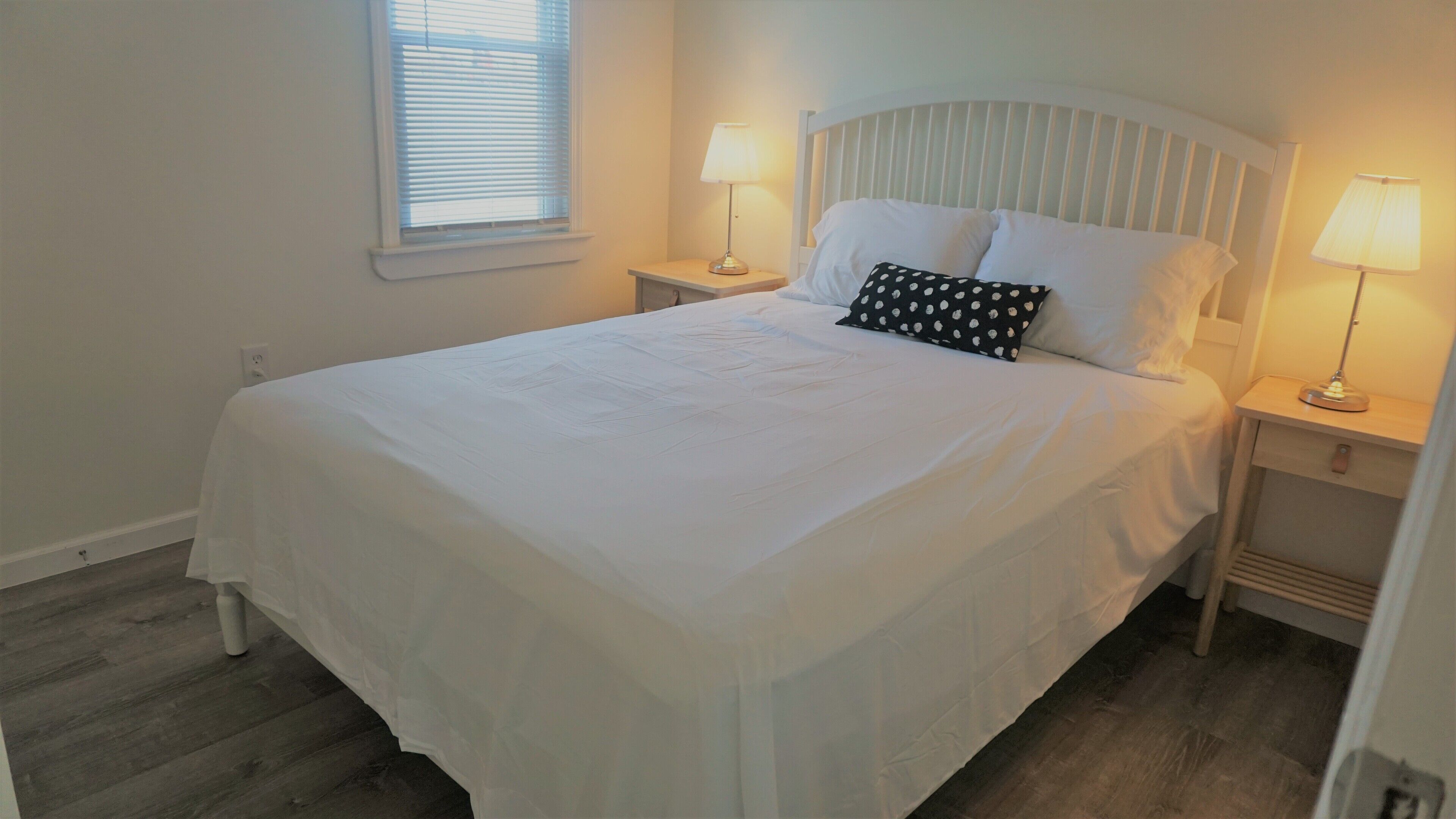 A neatly made bed with white linens, two bedside tables with lamps, and a window with blinds.