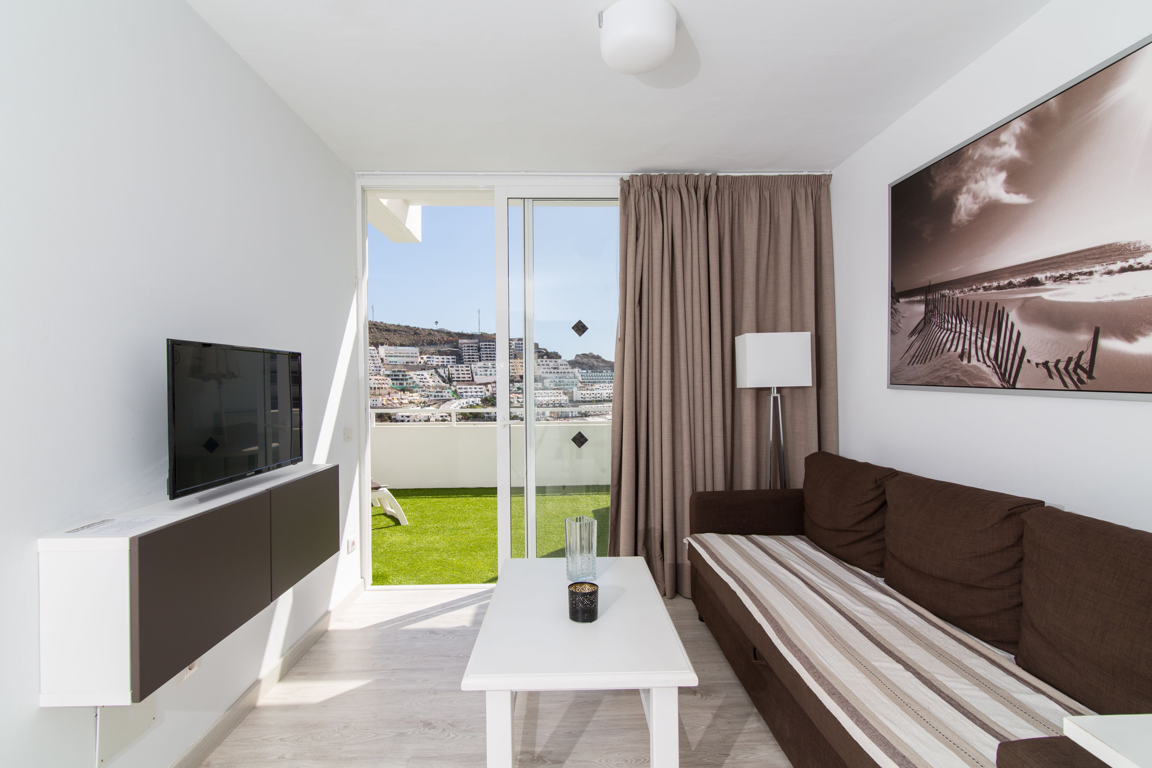 A modern living room with a TV, a sofa, a table, and a view of a cityscape through a glass door.