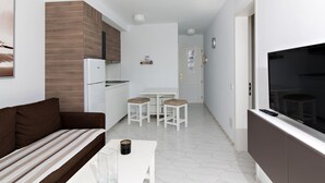 A modern kitchen with white cabinets, a built-in oven, and a refrigerator. There is a dining area with a white table and two stools. A TV is mounted on the wall, and a sofa is placed in the living area. - Apartamentos Los Mocanes (Mogan)