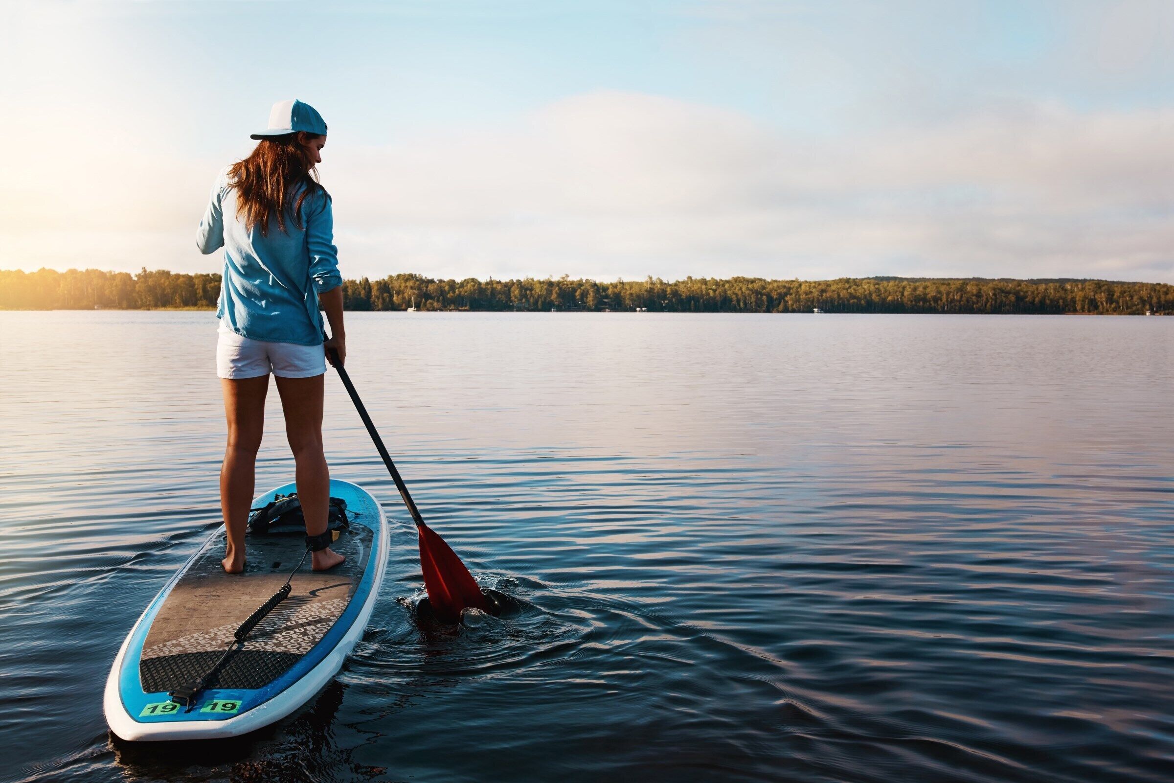 Eine Person steht auf einem Paddleboard auf einem ruhigen See, im Hintergrund eine bewaldete Uferlinie.