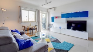 A modern living room with a sofa, a glass coffee table, a dining area with a table and chairs, a TV mounted on a white wall, and a balcony with a view. - Centric and Spacious Apartment With Wifi and Balcony (Gzira)