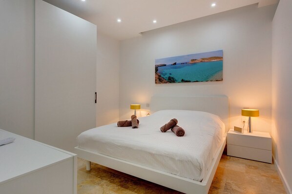 A modern bedroom with a large bed, two bedside lamps, and a painting of a coastal scene. - Modern 2 Bedroom Apartment in St Julians (St. Julian's)