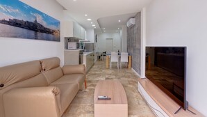 Ein modernes Wohnzimmer mit einer Couch, einem Couchtisch und einem Fernseher. Im Hintergrund ist eine Küche mit Essbereich zu sehen.